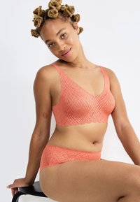Set di reggiseno e slip corallo con design testurizzato e patterned, con bordi ondulati e spalline larghe, mostrato su un modello seduto.