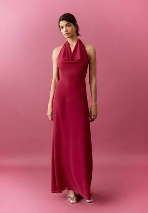 Giovane donna con capelli scuri in un abito rosso senza maniche, lungo fino a terra, e tacchi argento aperti sulle punte, in piedi davanti a uno sfondo rosa uniforme.