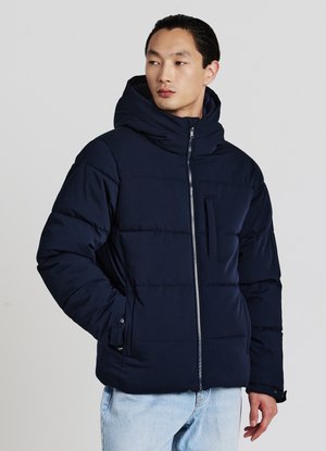 Giacca imbottita blu navy con cappuccio alto, chiusura frontale con zip e tasca sul petto. Presenta una texture in tessuto liscia e polsini elastici.