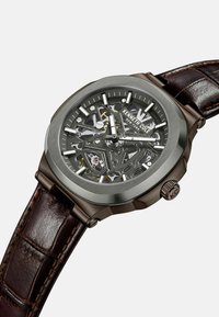 Zegarek typu skeleton z brązowym skórzanym paskiem, stalową kopertą i przezroczystą tarczą, ukazującą wewnętrzne mechanizmy. Srebrne wskaźniki godzinowe.