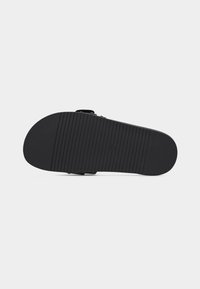 Semelle de sandale noire avec des rainures horizontales pour l'adhérence, vue par le dessous sur un fond clair.