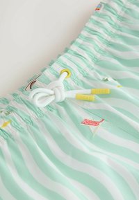 Hellgrün-weiß gestreifte Badeshorts mit weißem Kordelzug, der zu einem Knoten gebunden ist, gelben Enden mit der Aufschrift "NEXT" und kleinen bestickten Strandmotiven.