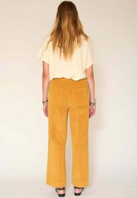 Pantalons en velours côtelé moutarde taille haute avec jambes droites et poches arrières, associés à un haut crème ample. Tissu texturé visible.