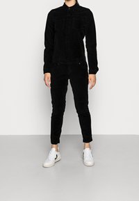 Combinaison en velours côtelé noire avec des manches longues, fermeture à boutons sur le devant et deux poches poitrine, portée avec des baskets blanches à accents noirs.