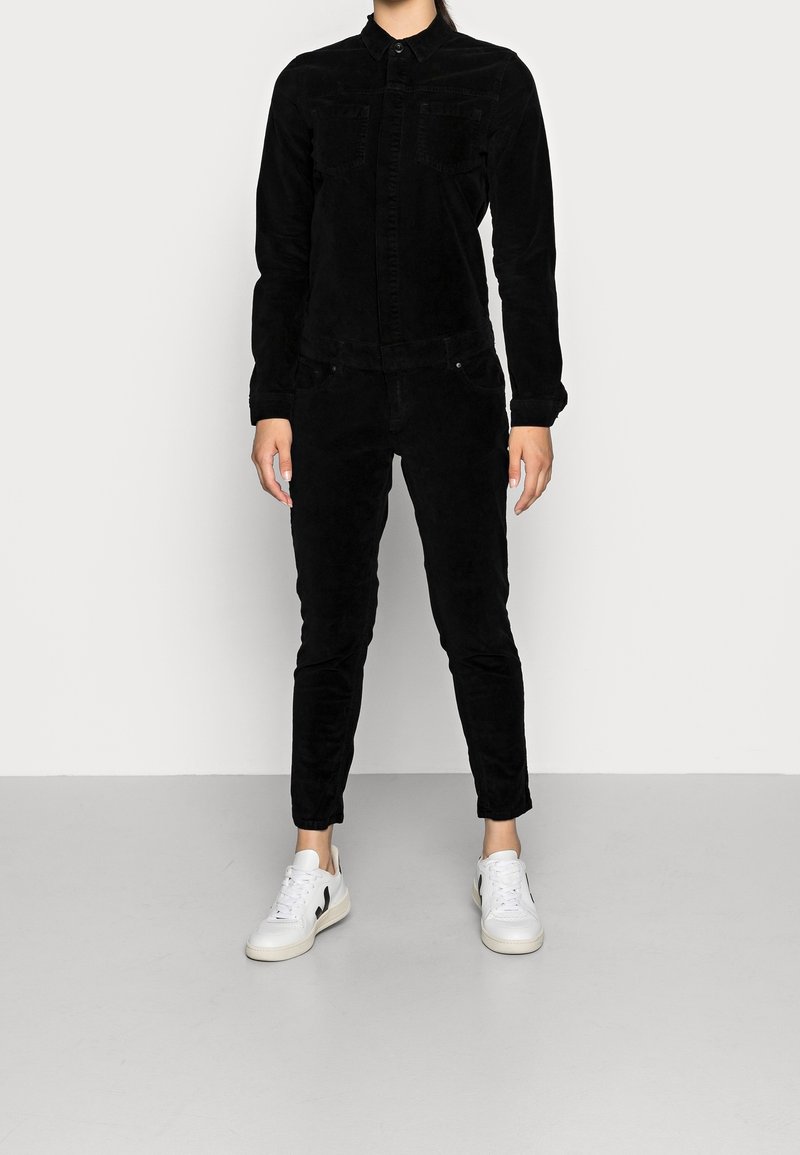 Combinaison en velours côtelé noire avec des manches longues, fermeture à boutons sur le devant et deux poches poitrine, portée avec des baskets blanches à accents noirs.