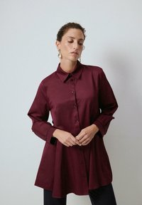 Woman wearing a long-sleeve burgundy button-up shirt and gold drop earrings, standing against a plain light gray background.