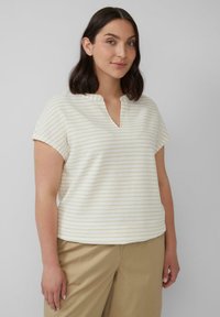 Kobieta z ramionami do ramion, mająca ciemne włosy, ubrana w krótką białą i beżową bluzkę w paski oraz beżowe spodnie, stojąca na jednolitym tle.