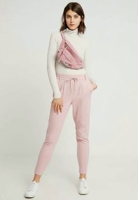 Γυναίκα όρθια, φορώντας λευκό ζιβάγκο, ροζ ανοιχτό παντελόνι φόρμας, λευκά αθλητικά παπούτσια και ροζ τσαντάκι μέσης διαγώνια.