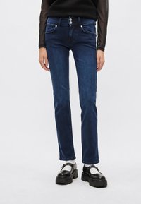 Jeans de mezclilla azul oscuro con un ajuste recto y ceñido, diseño de cinco bolsillos y herrajes plateados. Presentan un cierre de doble botón en la cintura.