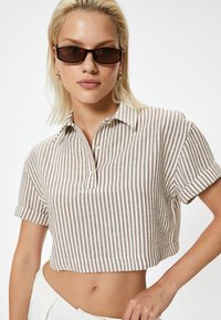 Camisa corta de manga corta con un patrón de rayas en beige y blanco, con cuello y un cierre de un solo botón en el cuello.