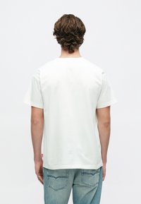 Λευκό βαμβακερό μπλουζάκι με κλασικό στρογγυλό λαιμό και κοντά μανίκια, με ίσια ραφή στο τελείωμα και μικρή κόκκινη ετικέτα στο πλάι.