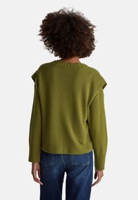 Zelený pletený svetr se žebrovaným kulatým výstřihem a lemem, s kleslými rameny a volným střihem. Nošený s modrými džínami.