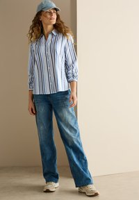 Camicia a righe blu chiaro e bianca, con un taglio rilassato, abbinata a jeans blu larghi e sneakers bianche, completata da un cappellino blu chiaro.