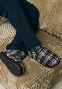 Sivé a čierne kárované papuče s chlpovým povrchom a čiernymi gumovými podrážkami, nosené s tmavozelenými nohavicami na ryhovanom béžovom podklade.