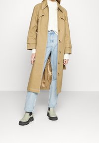 Tan trench coat with a glossy inner lining, paired with a white textured sweater, light blue jeans, and green ankle boots with black accents.