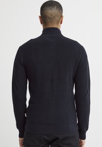 Marineblå strikket genser med høy krage, ribbestrikket tekstur og tettsittende design. Har lange ermer og en subtil merkeetikett på kanten.