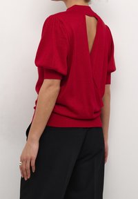 Haut en maille rouge avec manches ballon, décolleté en V dans le dos et taille cintrée. La texture présente un léger scintillement. Associé à un pantalon noir ajusté.