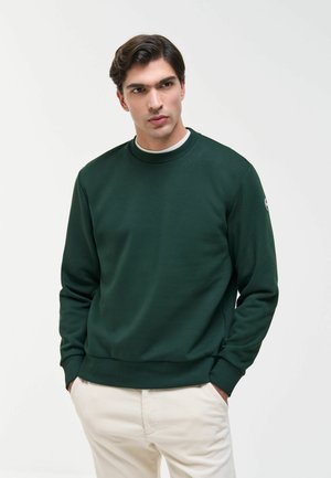 Uomo che indossa una felpa verde scuro a maniche lunghe, con le mani nelle tasche e pantaloni beige, in piedi davanti a uno sfondo chiaro uniforme.
