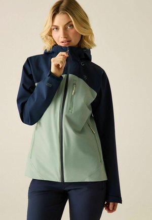 Blonde woman wearing a two-tone waterproof jacket, light green front and navy blue sleeves, zipping up the front zipper against neutral background.
