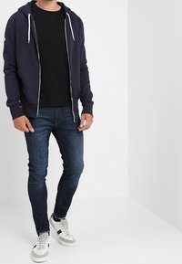 Námořnická mikina na zip s bílými šňůrkami, černé tričko, tmavé džíny a bílé tenisky se šedými detaily. Jednoduchý, neformální design.