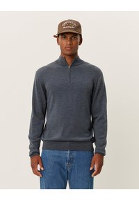 Pulover gri cu fermoar la gât, confecționat dintr-un material moale, având mâneci lungi și o bandă de jos ajustată. Purtați cu blugi din denim albastri. Căciulă cu logo pe cap.