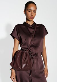 Braune Leder-Eimer-Handtasche mit gerafftem Oberteil und Tragegurt, kombiniert mit einem passenden braunen strukturierten Hemdkleid mit kurzen Ärmeln und Knopfverschluss.