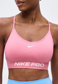Γυναίκα φοράει ένα ροζ αθλητικό σουτιέν Nike Pro με λεπτές τιράντες και λευκό λογότυπο Nike, συνδυασμένο με ανοιχτό μπλε αθλητικό κάτω μέρος.