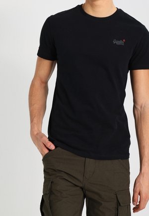 Homme portant un t-shirt noir uni à manches courtes et un pantalon cargo vert olive, une main dans la poche, sur un fond blanc.