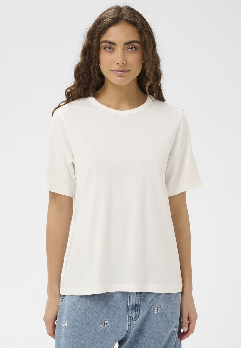 Femme aux longs cheveux bouclés portant un T-shirt blanc à manches courtes uni et un jean bleu clair brodé, debout sur un fond blanc.