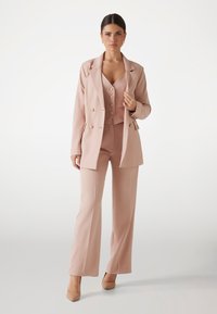 Helles rosa maßgeschneidertes Anzug mit einem doppelt geknöpften Blazer, passender taillierter Weste mit Knöpfen und weit geschnittenen Hosen, kombiniert mit hautfarbenen High Heels.