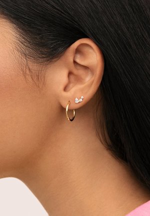 Pendiente de aro dorado con un pequeño diseño que presenta dos acentos de piedras preciosas transparentes, una en forma de pera y otra redonda, colocadas cerca del lóbulo de la oreja.