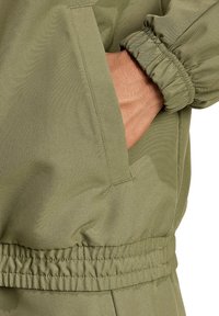 Olivgrüne Jacke mit elastischen Bündchen und Taillengummi, versehen mit einer seitlichen Taschenverzierung. Glattes, leichtes Material mit matter Oberfläche.