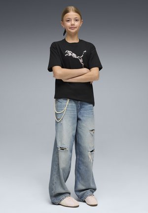 Dziewczyna z założonymi na siebie rękami, ubrana w czarną koszulkę z nadrukiem, podarte szerokie jeansy z perłowym łańcuszkiem oraz białe sneakersy na szarym tle.