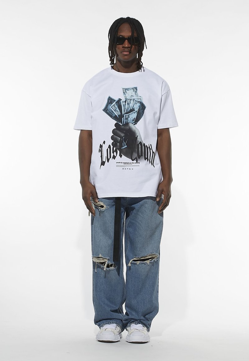 Fehér, rövid ujjú póló, amelyen egy pénzt tartó kéz grafikája látható, kék farmernadrággal és fehér sportcipővel párosítva.
