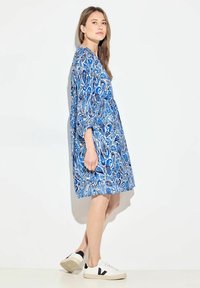 Vestido azul con estampado paisley, de manga tres cuartos, corte hasta la rodilla y un ajuste relajado, combinado con zapatillas blancas con detalles en negro.
