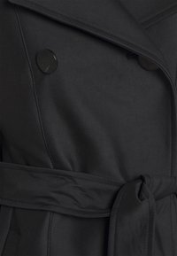 Černý trench coat s dvojitým zapínáním, hladkou tkaninou, kulatými černými knoflíky a zavazovacím páskem v pase.