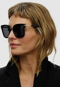 Sorte oversized solbriller med en kraftig rektangulær ramme, guldaccentuer på tempelene, mørke linser og en blank overflade.