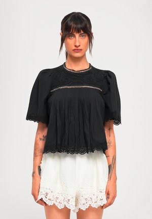 Vrouw met donker haar in een zwarte blouse met kant en witte geborduurde shorts, staande tegen een effen lichte achtergrond.