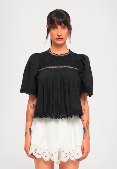 Femme aux cheveux bruns portant un blouse noire à bordures en dentelle et un short blanc brodé, se tenant devant un fond léger uni.