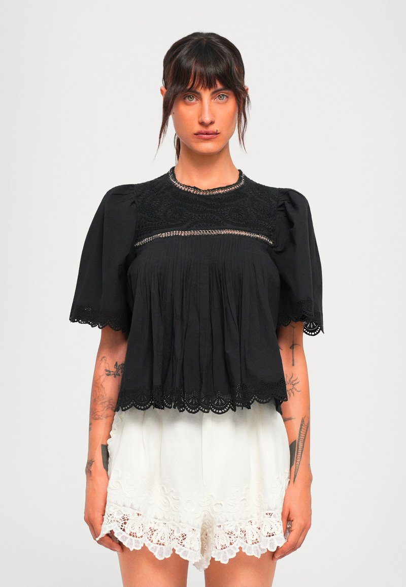 Woman with dark hair in a black lace-trimmed blouse and white embroidered shorts, standing against a plain light background.