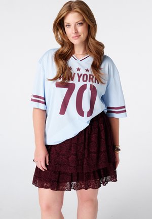 Femme aux longs cheveux châtain clair portant un maillot de sport oversize bleu clair New York 70 et une jupe en dentelle rouge foncé, debout sur un fond uni.