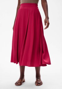 Lange, geplooide rok van vibrante magenta stof, met een hoge tailleband en een zachte, vloeiende textuur. Draagt met bruine, gespsandalen.