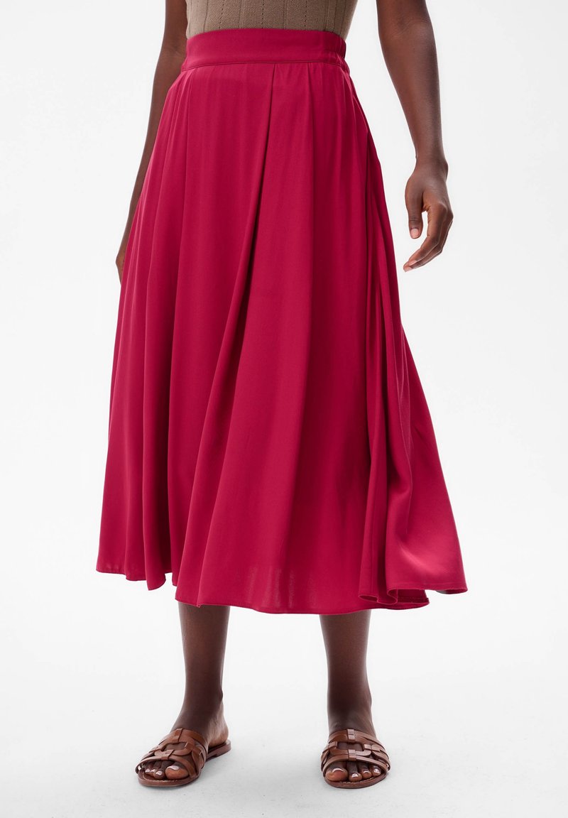 Lange, geplooide rok van vibrante magenta stof, met een hoge tailleband en een zachte, vloeiende textuur. Draagt met bruine, gespsandalen.
