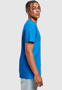 Blå bomulds t-shirt med korte ærmer og rund hals. Har grafisk tryk på forsiden. Modellen står sideways iført denim jeans.