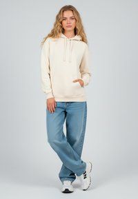 Felpa beige con tasca frontale, coulisse e polsini a coste, abbinata a jeans azzurri a vestibilità rilassata e scarpe da ginnastica bianche.