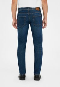 Jeans de mezclilla azul oscuro con un corte recto, que presentan un parche de cuero marrón con el logo, dos bolsillos traseros y detalles de costura sutiles.