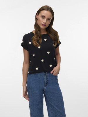 Vrouw met lang bruin haar die een zwart T-shirt met korte mouwen met witte hartjesprint en een blauwe high-waisted jeans draagt, staand tegen een witte achtergrond.