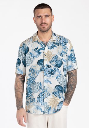 Mężczyzna z krótkimi włosami i tatuażami, ubrany w niebieską, beżową i białą kwiecistą koszulę na krótkim rękawie zapinaną na guziki oraz jasne spodnie.