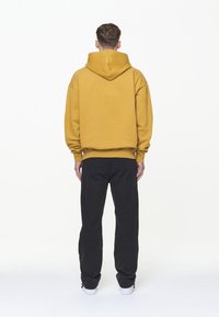 Felpa con cappuccio di colore giallo senape, caratterizzata da una vestibilità rilassata e polsini a coste, abbinata a pantaloni neri, vista da dietro.