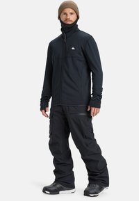 Giacca in fleece nera con zip e dettagli a pannello, abbinata a pantaloni da neve neri larghi e stivali da neve neri, tutti adatti per attività invernali.